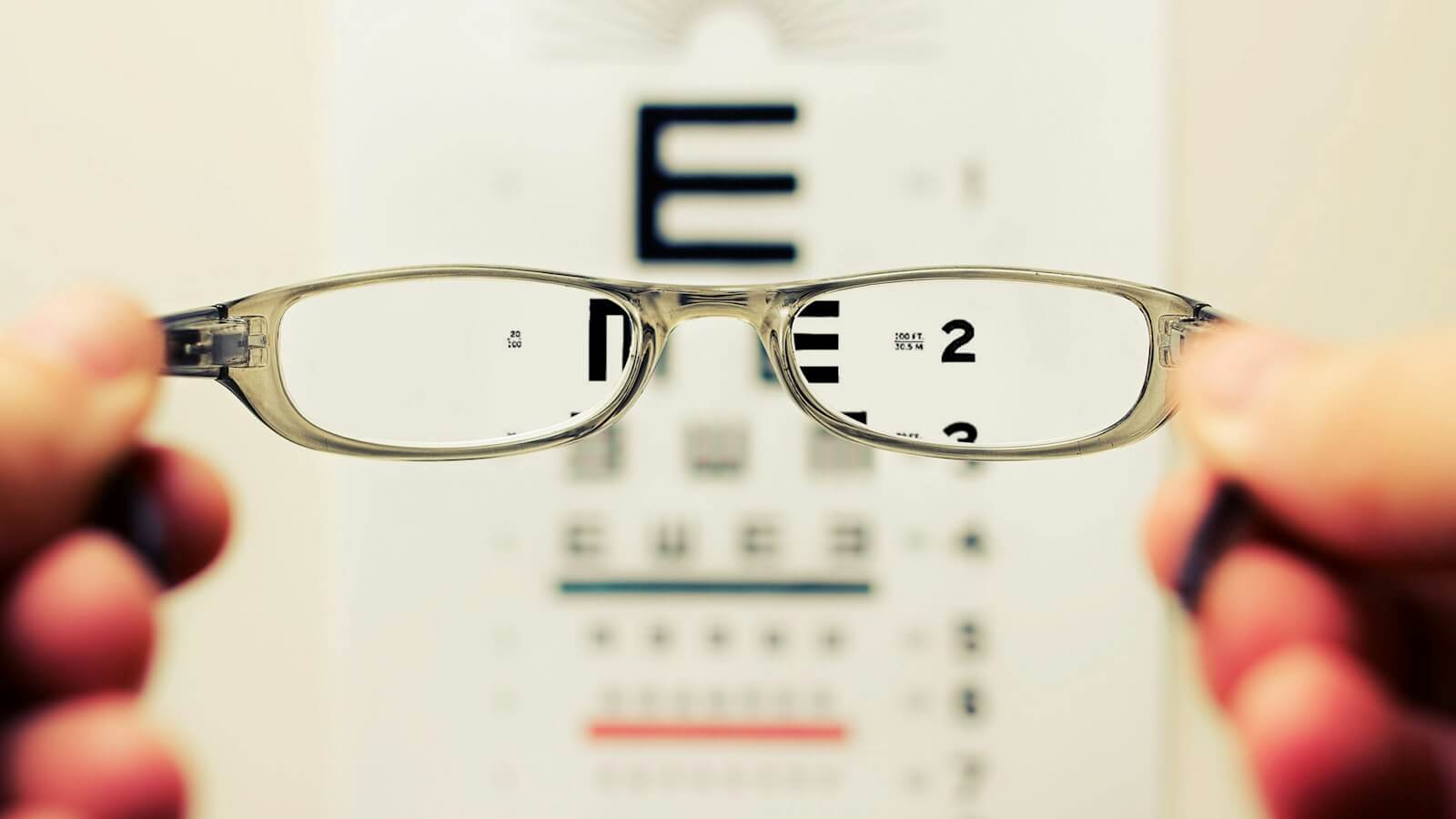 Hands holding reading glasses that bring a blurry eye chart into focus